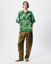 A model stands against a light gray studio background wearing a green short-sleeve button-up shirt with a dark abstract print and loose olive-brown pants.