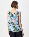 A woman shown from the back wearing a sleeveless V-neck blouse with a colorful floral print in shades of blue, pink, green, and white, paired with dark trousers against a plain white studio background.