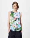 A woman modeling a sleeveless top with a vibrant watercolor floral print in shades of green, white, pink, and blue, paired with dark navy trousers, against a plain light gray background.