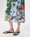 A person wearing a knee-length floral skirt with large white, blue, pink, and green flowers over a light background, paired with black flip-flop sandals, photographed against a clean white studio backdrop.