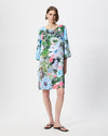 A woman modeling a knee-length, three-quarter sleeve dress with a vibrant floral print in shades of blue, pink, green, and white, paired with black flip-flop sandals against a plain white studio background.