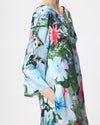 A close-up of a model wearing a loose, short-sleeved kaftan dress with a vibrant watercolor floral print in shades of blue, green, pink, and white against a light gray background.