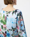 A close-up back view of a woman wearing a long-sleeved blouse with a vibrant watercolor floral print in shades of blue, green, pink, and white.