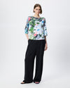 A model stands against a light gray studio background wearing a floral print top with three-quarter sleeves and black wide-leg trousers.