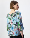 A woman shown from the back wearing a short-sleeved blouse with a vibrant floral print in shades of blue, green, pink, and white over a dark base, paired with black trousers against a plain white studio background.