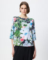 A woman modeling a short-sleeved blouse with a vibrant floral and botanical print in shades of green, blue, pink, and white, paired with black trousers against a plain light gray background.