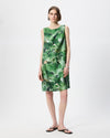 A woman modeling a sleeveless knee-length dress with an abstract green and white watercolor-style print, paired with black flip-flop sandals against a plain light gray studio background.