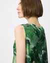 A woman shown from the side and back wearing a sleeveless green top with an abstract watercolor-style print in various shades of green, black, and white, against a plain light gray background.
