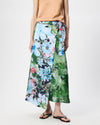 A model wearing a green and blue floral wrap maxi skirt with a tie waist, paired with a tan top and black flip-flops, against a plain white studio background.