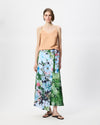 A model stands against a light gray studio background wearing a peach-colored spaghetti-strap camisole top and a long, wide-leg wrap skirt featuring a vibrant blue, green, and pink floral print.