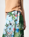 A close-up of a model wearing a wrap skirt with a split floral print design, featuring light blue flowers on one side and a green botanical print on the other, paired with a peach-colored top against a light gray background.