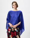 A woman modeling a royal blue satin-like draped top with wide, flowing sleeves, paired with black floral pants, against a plain light gray studio background.