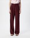 A model wearing a pair of high-waisted, wide-leg trousers in a deep burgundy color, paired with a light pink top and black sandals, against a plain white studio background.