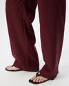 Close-up of a person's legs wearing wide-leg burgundy pants and dark brown flat thong sandals against a plain white studio background.