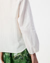 Close-up of a person wearing a white textured long-sleeve blouse with sheer puff sleeves and green patterned bottoms, against a plain light background.