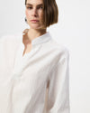 A close-up of a model wearing a white textured long-sleeve blouse with a high folded collar, set against a plain light gray studio background.