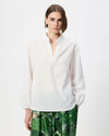 A woman modeling a white long-sleeve blouse with a stand collar and subtle vertical texture, paired with green patterned pants, against a plain light gray studio background.