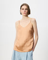 A woman modeling a light peach-colored spaghetti strap camisole top with a V-neckline, paired with light blue trousers, against a plain white studio background.