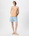 A model stands against a light gray studio background wearing a peach-colored spaghetti-strap camisole top, light blue denim shorts, and beige flip-flop sandals.