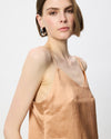 A woman wearing a light peach-beige satin camisole top with thin straps and a small chest pocket, photographed against a clean light gray studio background.