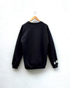 A black crewneck sweatshirt hanging on a wooden hanger against a white wall, featuring small embroidered flowers on the left sleeve cuff.