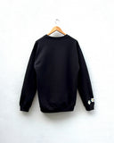 A black crewneck sweatshirt hanging on a wooden hanger against a white wall, featuring small embroidered flowers on the left sleeve cuff.