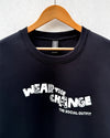 A black crewneck sweatshirt hanging on a wooden hanger against a light gray textured wall. The sweatshirt features a white graphic print on the front that reads 'WEAR THE CHANGE' with 'THE SOCIAL OUTFIT' printed underneath.
