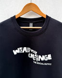 A black crewneck sweatshirt hanging on a wooden hanger against a light gray textured wall. The sweatshirt features a white graphic print on the front that reads 'WEAR THE CHANGE' with 'THE SOCIAL OUTFIT' printed underneath.