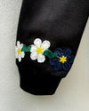 Close-up of a black sleeve with embroidered flowers along the cuff, featuring white and navy blue blossoms with yellow centers and green stems, against a light textured wall.