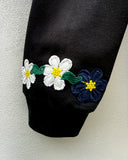 Close-up of a black sleeve with embroidered flowers along the cuff, featuring white and navy blue blossoms with yellow centers and green stems, against a light textured wall.