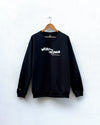 A black crewneck sweatshirt hanging on a wooden hanger against a white textured wall. The sweatshirt features a white graphic text print on the chest reading 'WEAR THE CHANGE' with smaller text below.