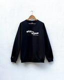 A black crewneck sweatshirt hanging on a wooden hanger against a white textured wall. The sweatshirt features a white graphic text print on the chest reading 'WEAR THE CHANGE' with smaller text below.