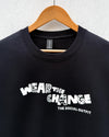 A black crewneck sweatshirt hanging on a wooden hanger against a light gray textured wall. The sweatshirt features a white graphic text print reading 'WEAR THE CHANGE' with a patterned fill and 'THE SOCIAL OUTFIT' underneath.