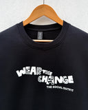 A black crewneck sweatshirt hanging on a wooden hanger against a light gray textured wall. The sweatshirt features a white graphic text print reading 'WEAR THE CHANGE' with a patterned fill and 'THE SOCIAL OUTFIT' underneath.