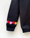 Close-up of a black ribbed sweatshirt sleeve with embroidered cherry blossom flowers in pink, red, yellow, and white near the cuff, shown against a light textured wall.