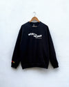 A black crewneck sweatshirt hanging on a wooden hanger against a white textured wall. The sweatshirt features a white graphic text print on the chest reading 'WEAR THE CHANGE' with smaller text below, and a small red and yellow graphic on the left sleeve cuff.