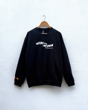 A black crewneck sweatshirt hanging on a wooden hanger against a white textured wall. The sweatshirt features a white graphic text print on the chest reading 'WEAR THE CHANGE' with smaller text below, and a small red and yellow graphic on the left sleeve cuff.
