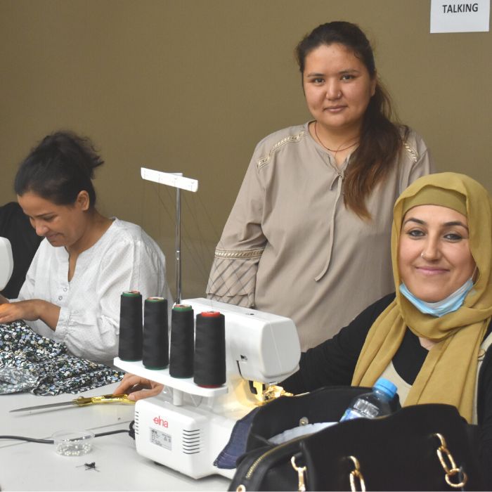 3 afghan women participating in sewing circle program learning to sew sitting at overlocking machine against green backdrop