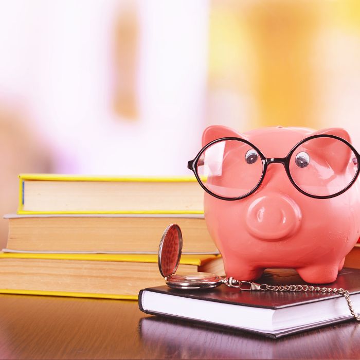 piggy bank wearing glasses with a pocket watch on top of financial literacy books on top of wooden table