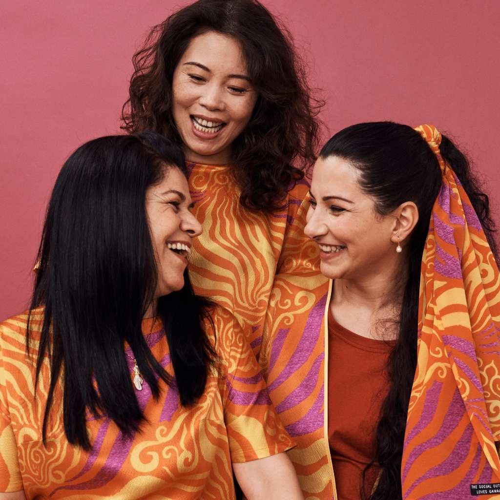 3 women with dark hair dressed in bright orange silk garments smiling at each other against a pink backdrop