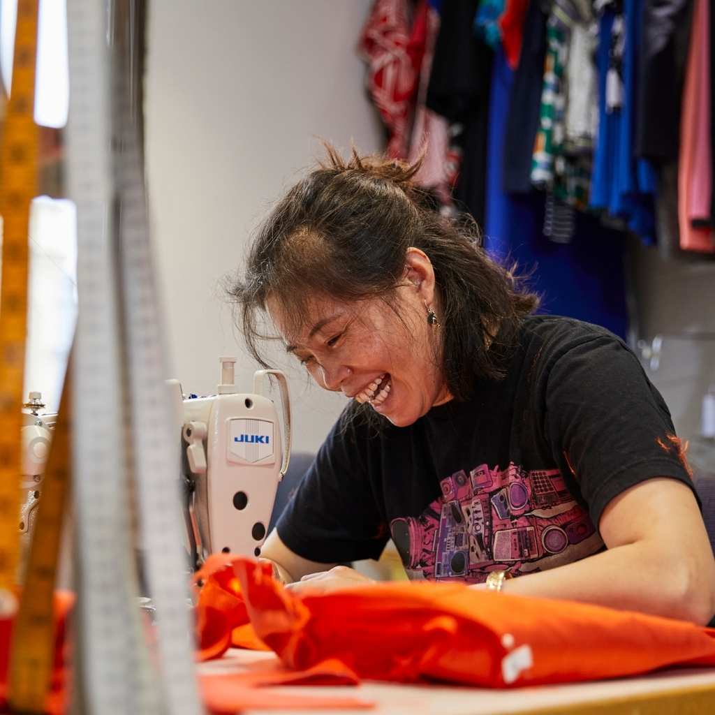 asian woman with long hair tied up in a ponytail sitting at sewing machine smiling and laughing while sewing, with measuring tapes in the foreground and colourful clothes in the backdrop