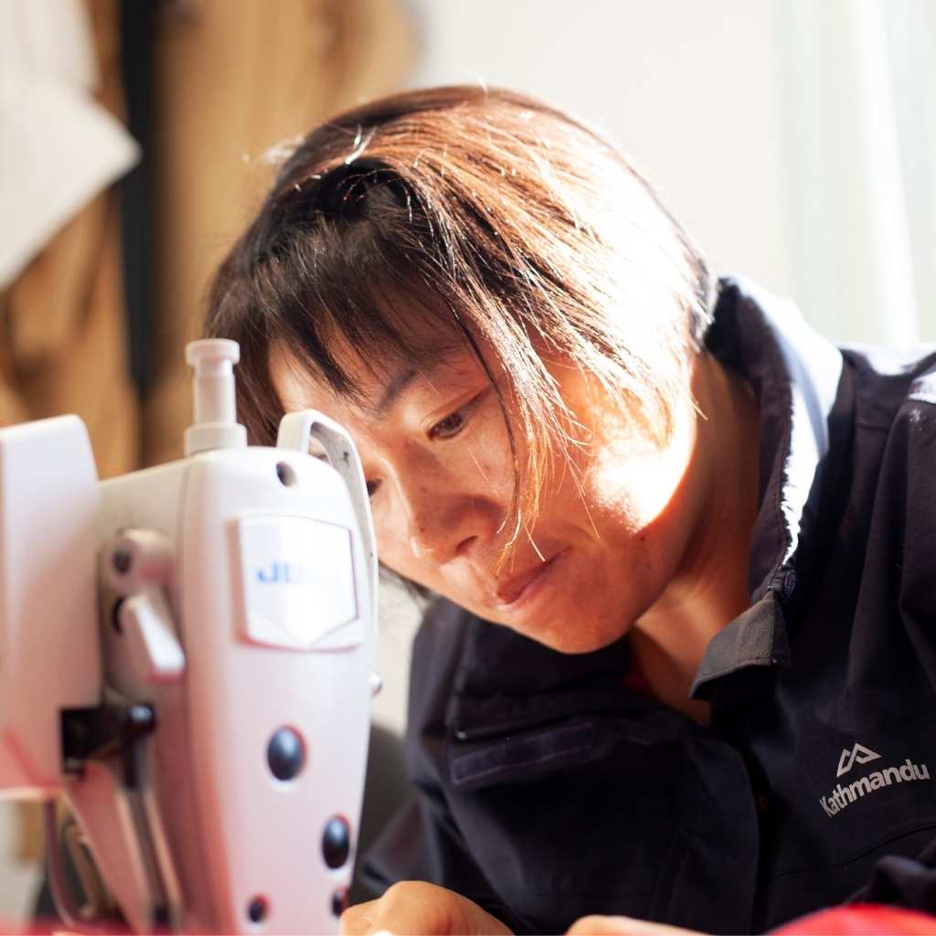 asian woman with short hair wearing a black jacket sitting at a sewing machine focusing on a task with the sun shining against her hair