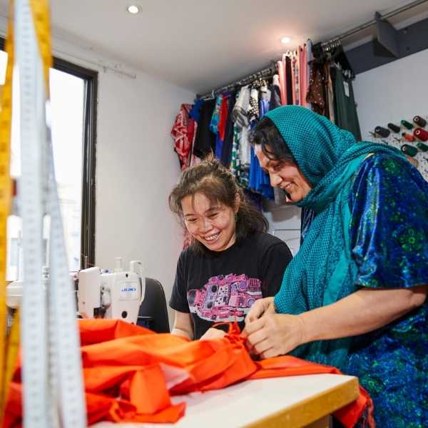 two women smiling and working with orange fabric in front of a sewing machine against a backdrop of colourful clothes and sewing bobbins