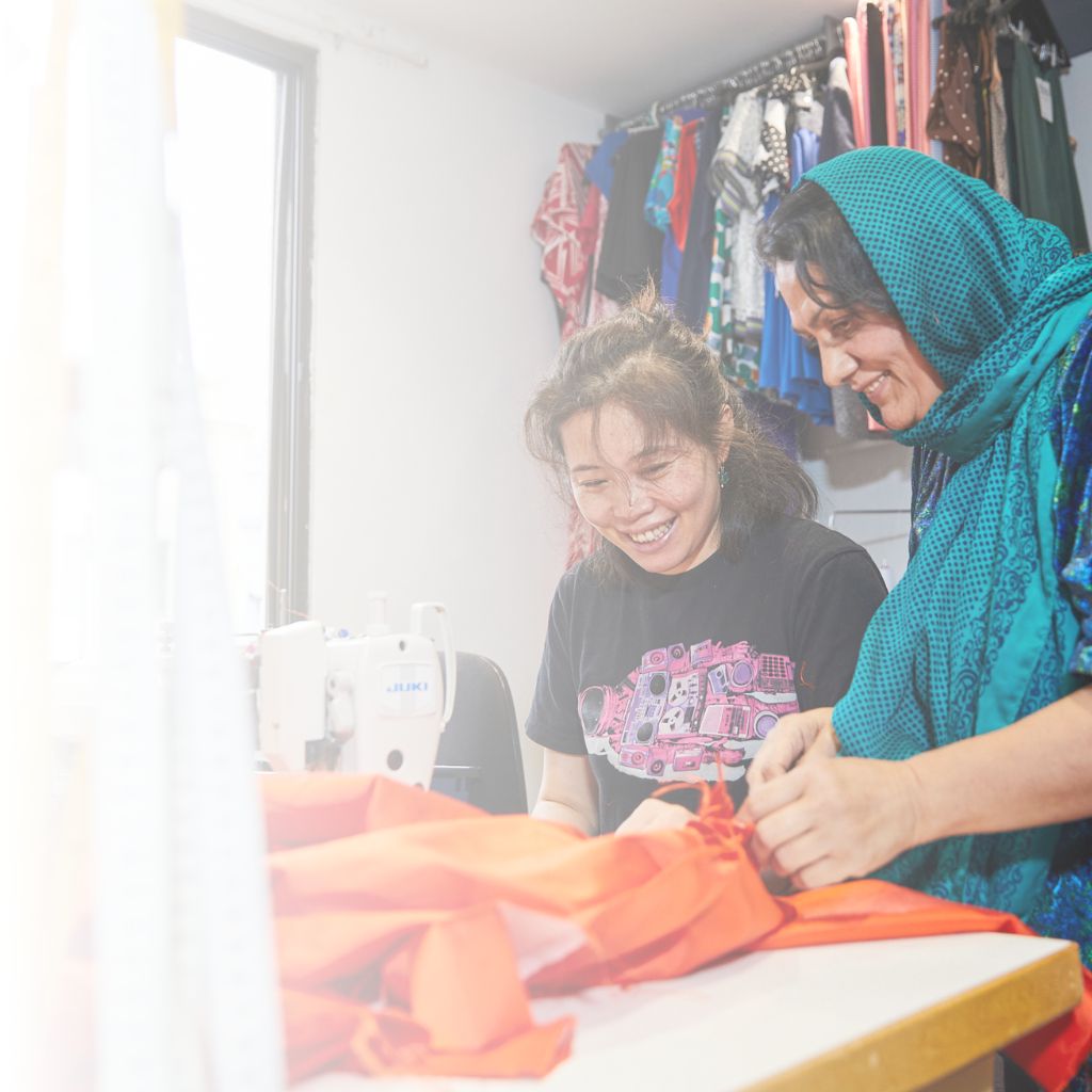 two women at sewing machine working on orange fabric together in the social outfit workroom