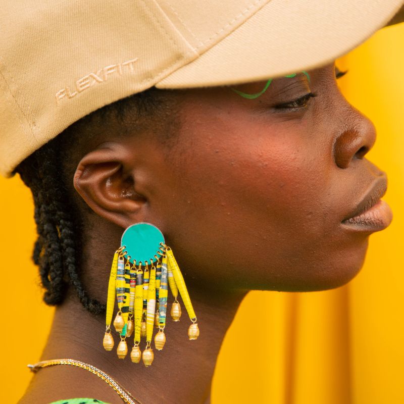 woman wearing tan cap with yellow dangly quazi earrings against yellow backdrop