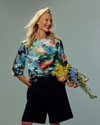 A smiling blonde woman stands against a muted blue-gray studio background, wearing a colorful floral three-quarter sleeve top and dark shorts while holding a bouquet of yellow, pink, and blue flowers.