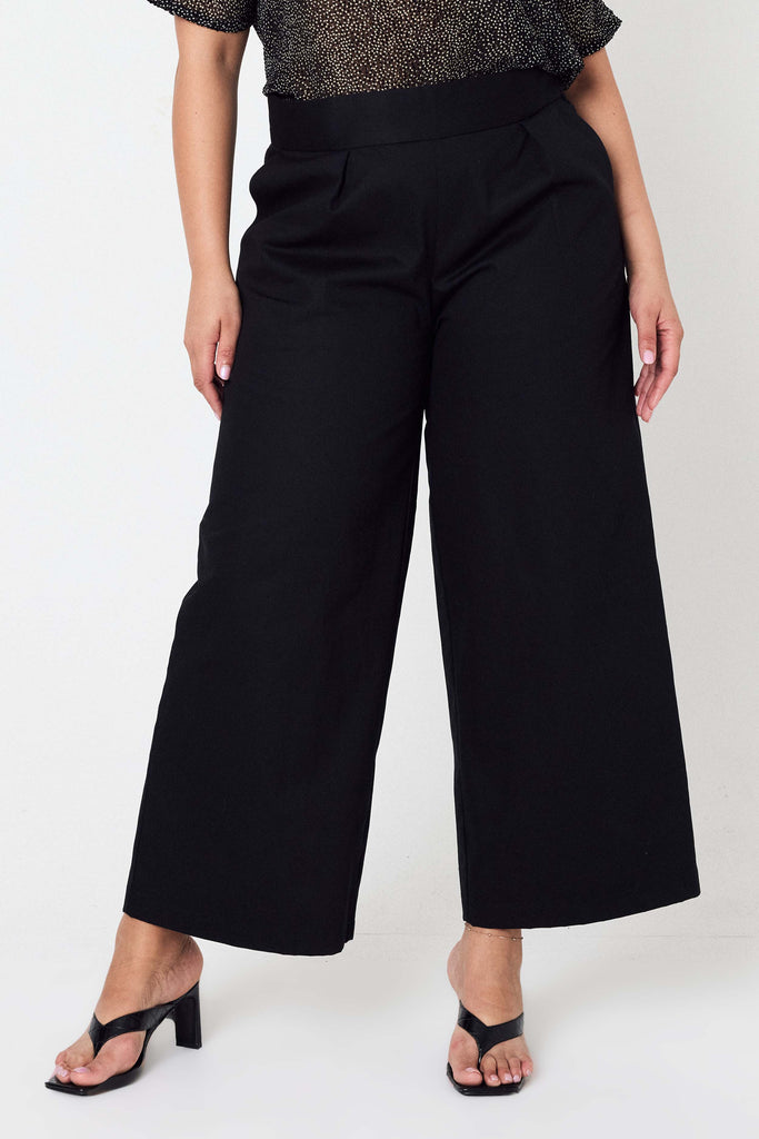 woman wearing black denim wide leg culottes, a sheer top and black heeled sandals standing against a white backdrop