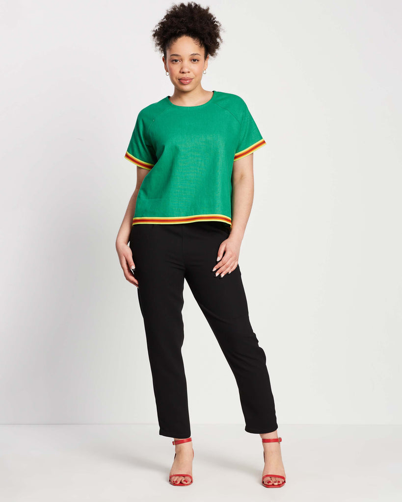 woman wearing green raglan sleeve top with contrast yellow and orange elastic ribbing on top of black straight leg pants with red strap heels