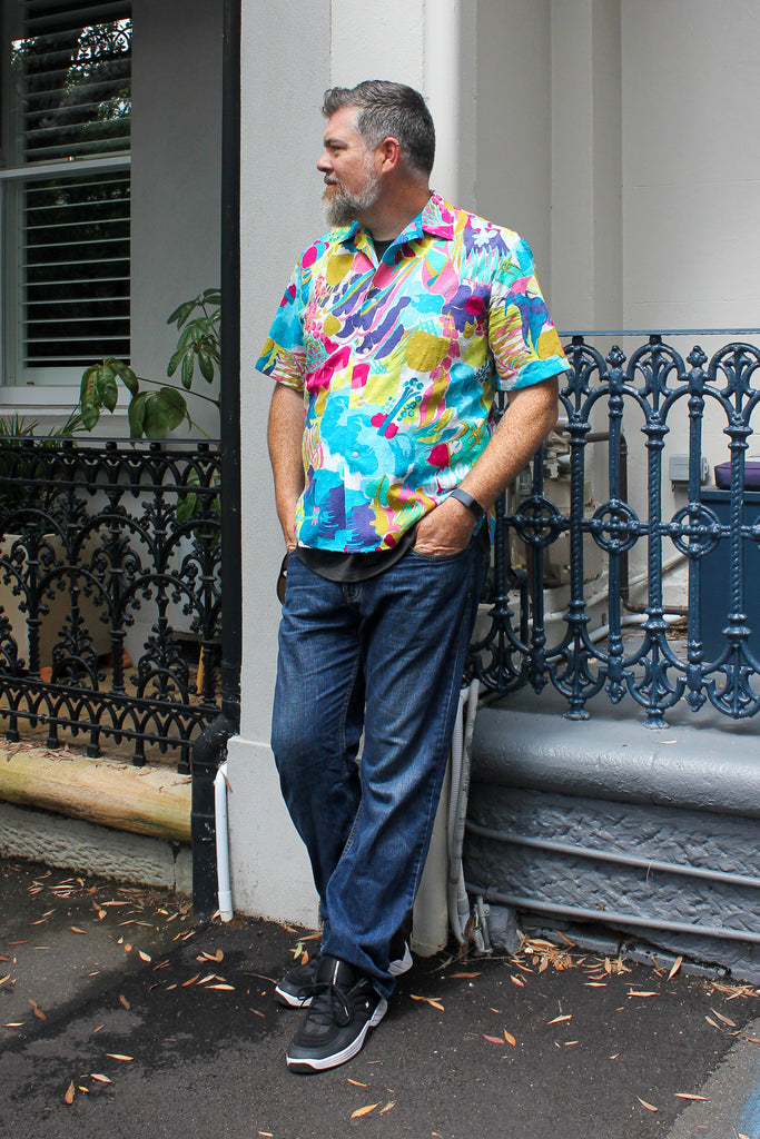 man wearing colourful floral shirt leaning against fence with blue jeans and black shoes