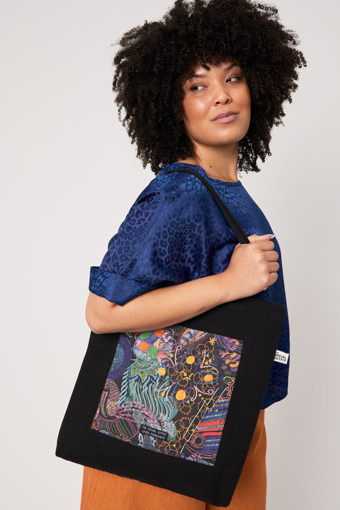 woman with curly hair looking away from the camera carrying a black tote bag wearing a blue top and orange linen pants against a white backdrop